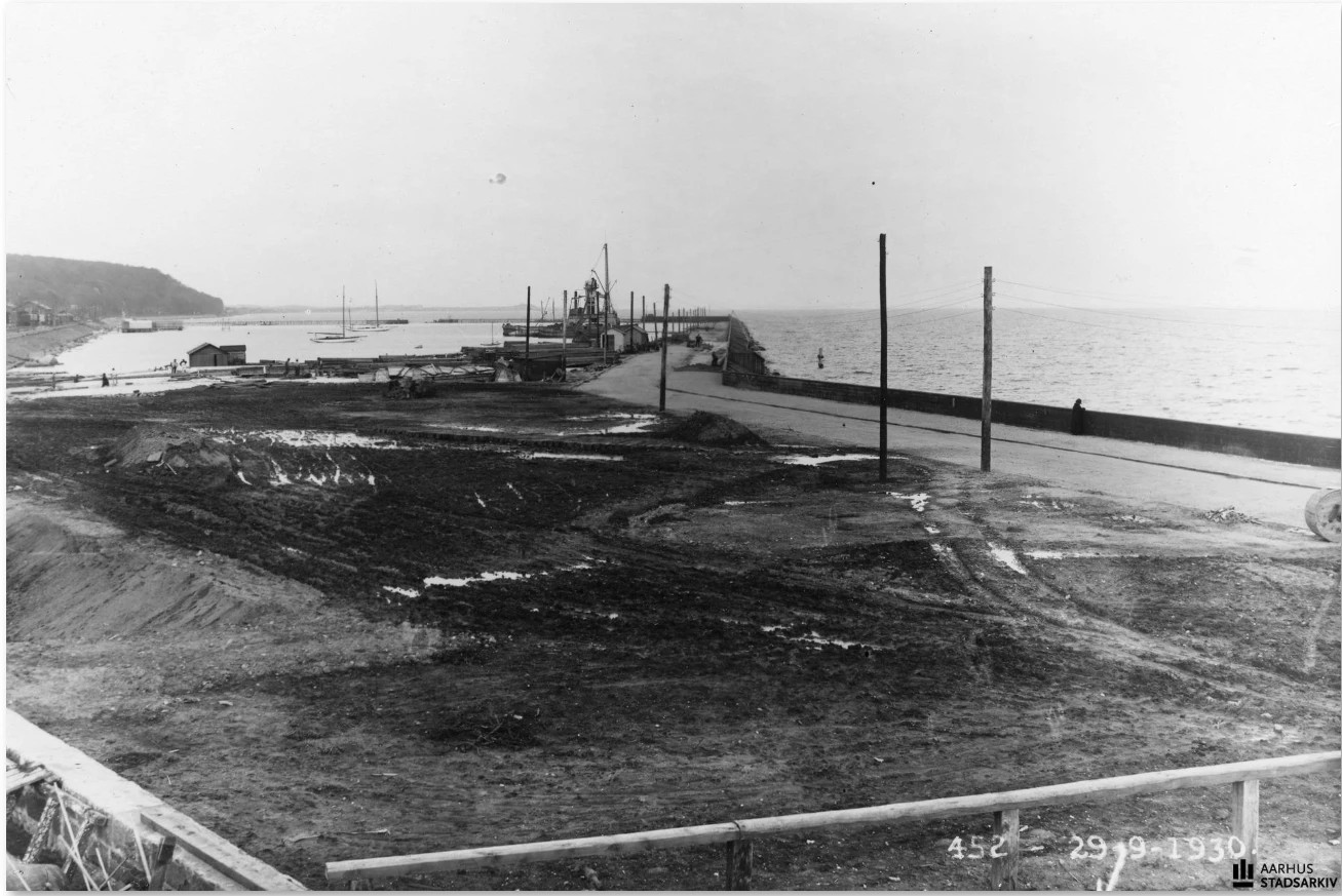 Sort-hvidt foto fra 1930 af Aarhus Havn under udvidelse: en byggeplads med mudret jord i forgrunden, en mole med vej til højre og sejlbåde og bygninger langs havnen i baggrunden.