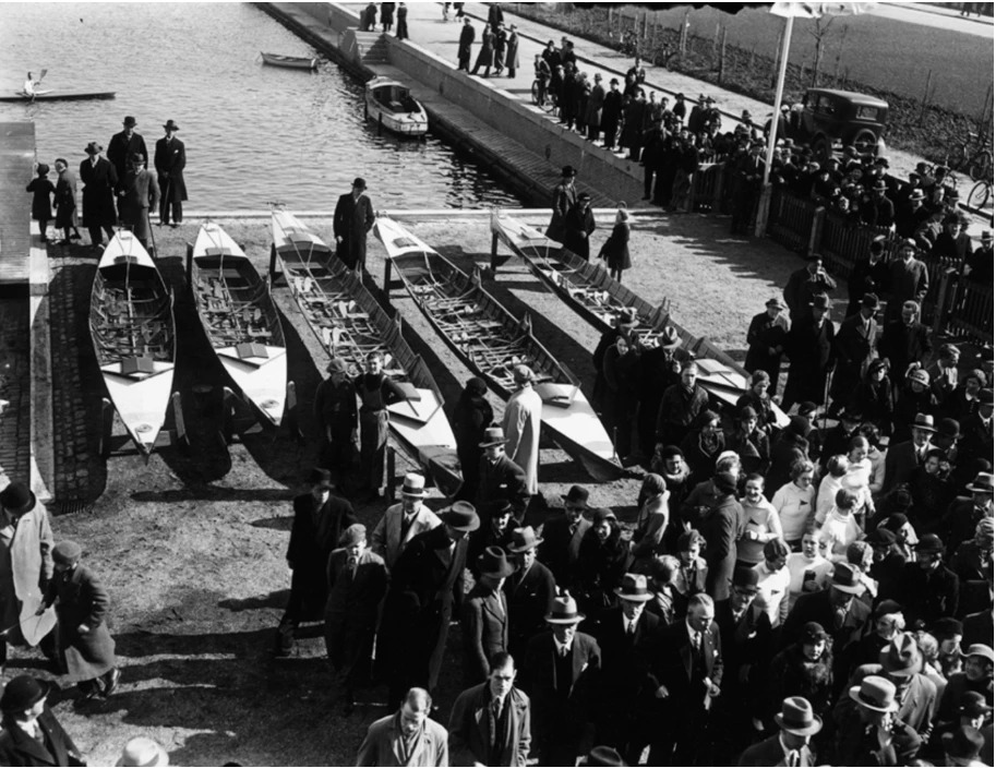 Sort-hvidt foto fra 1935 af fem både på land ved vandkanten, omgivet af en stor menneskemængde i hatte og frakker, med havnebassin og en bil i baggrunden.